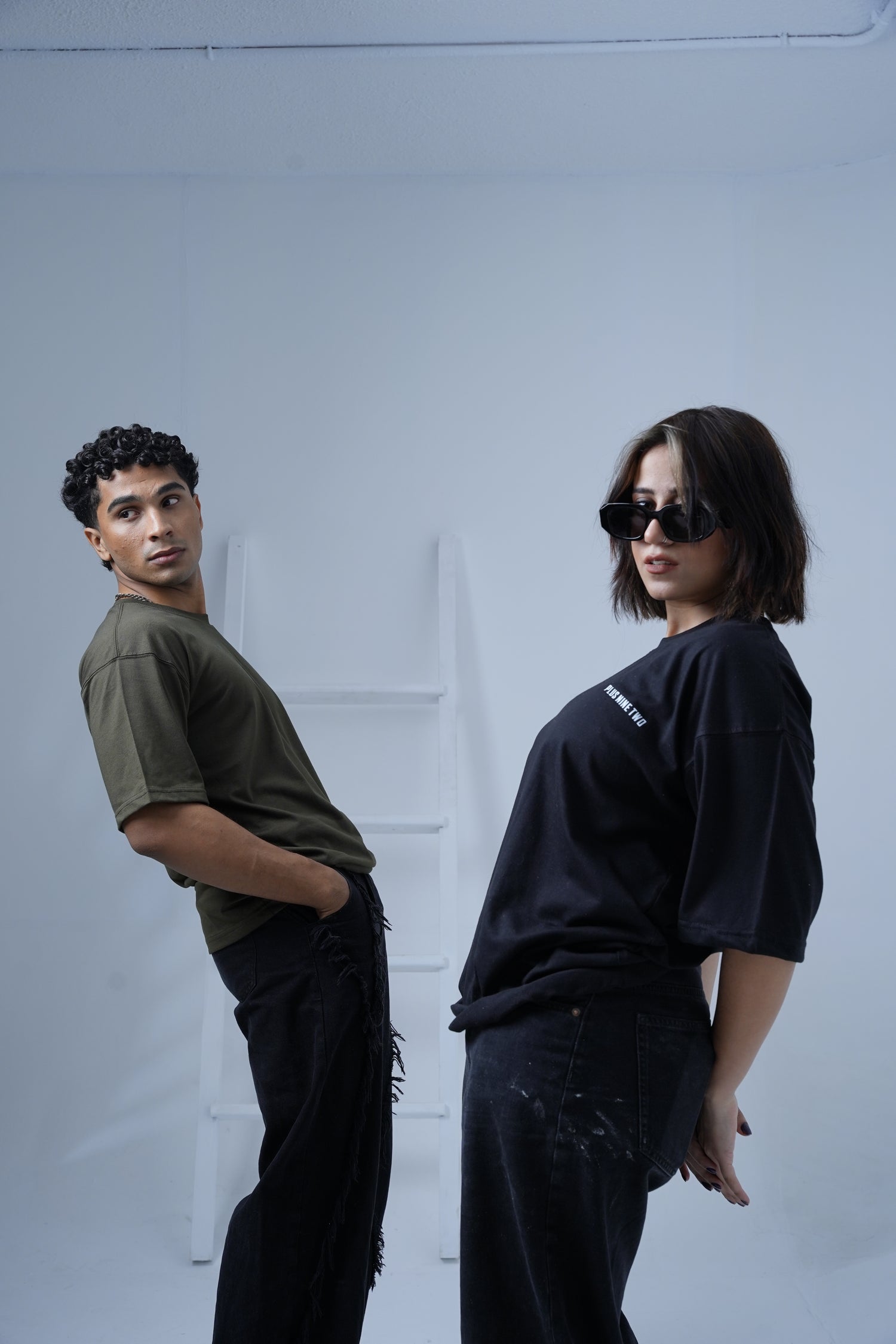 Two people standing on a white floor with a light blue wall wearing plus92 Essential Dropshirt in Army Green and Black