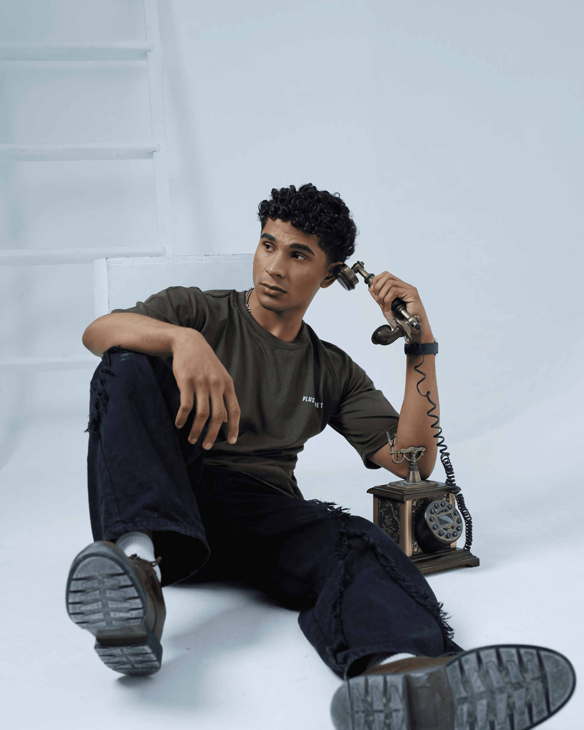 Person sitting on the floor wearing army green oversized t-shirt holding a vintage telephone against a light background