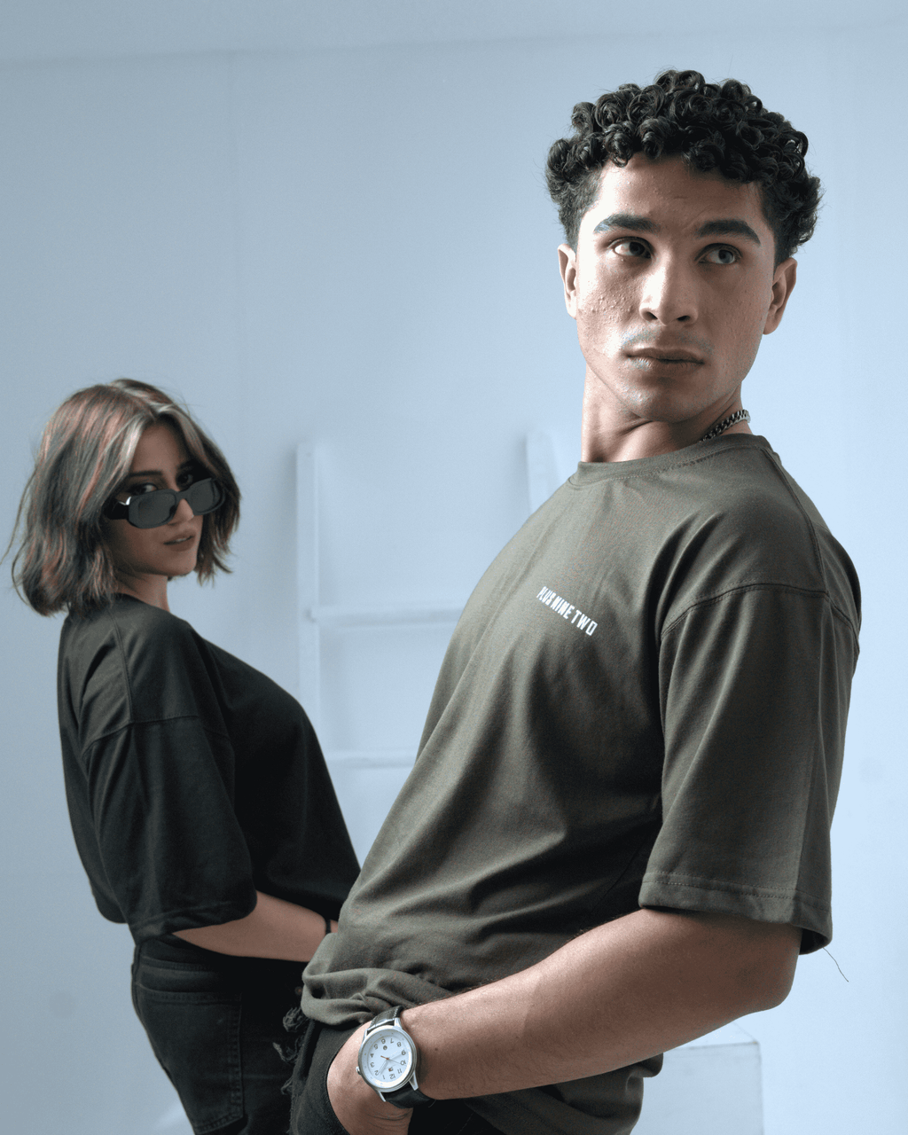Two people a man and a woman wearing oversized t-shirts with a brand logo against a light background