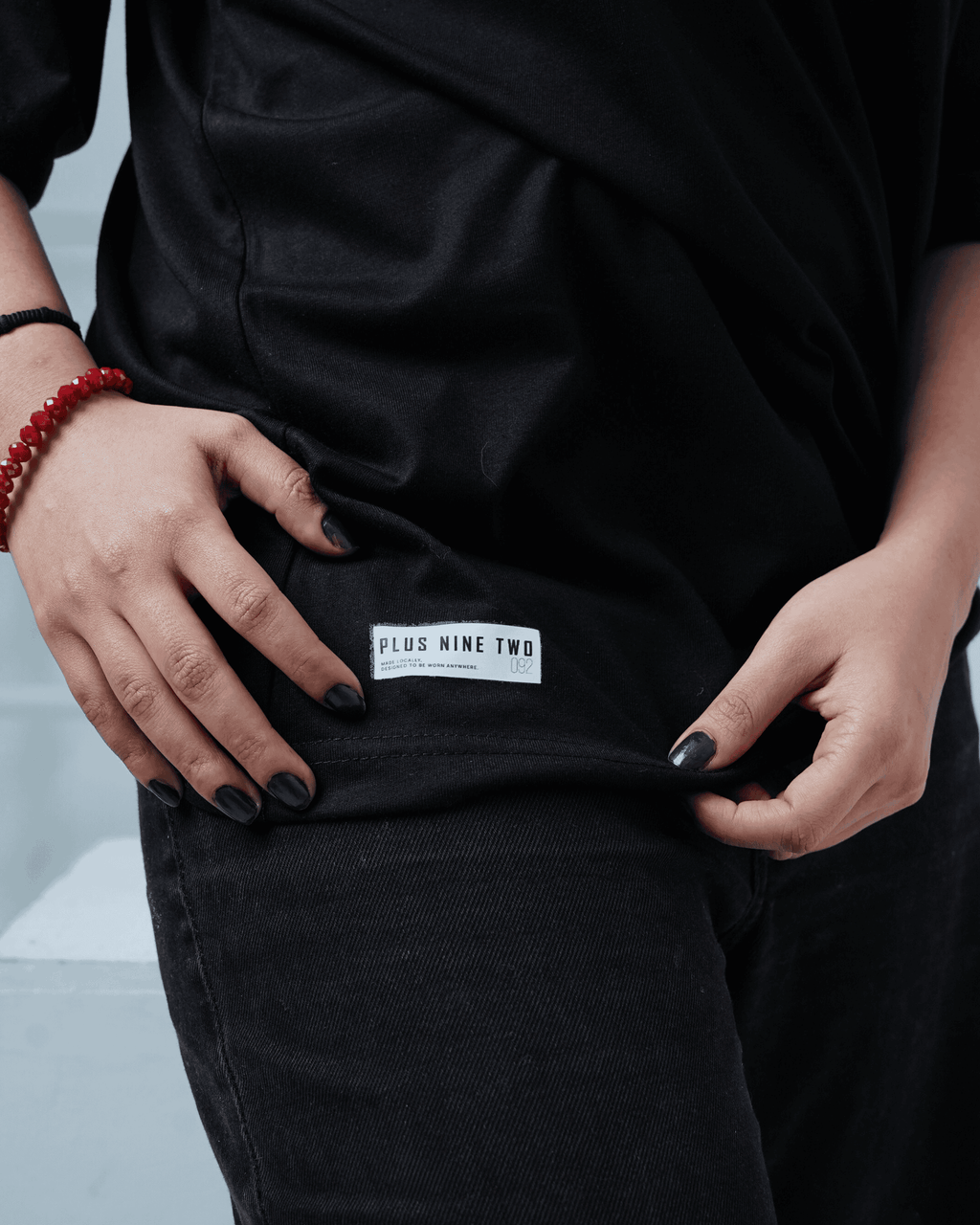 Person wearing a black oversized t-shirt with a visible +92clothing brand label on a light gray background