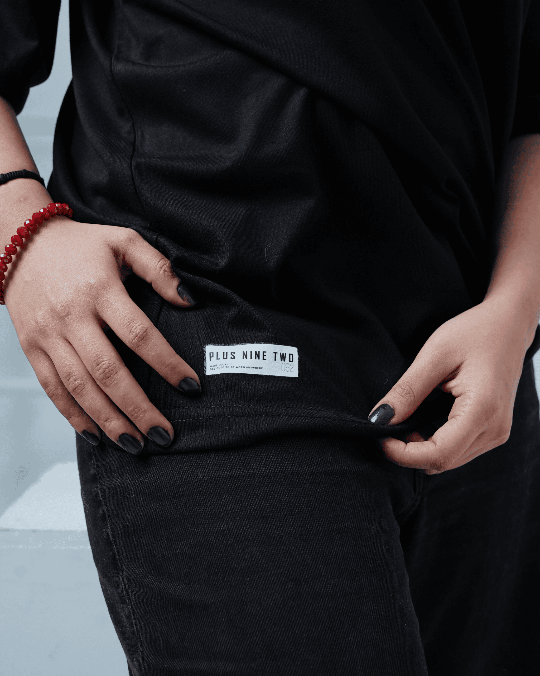 Person wearing a black oversized t-shirt with a visible +92clothing brand label on a light gray background