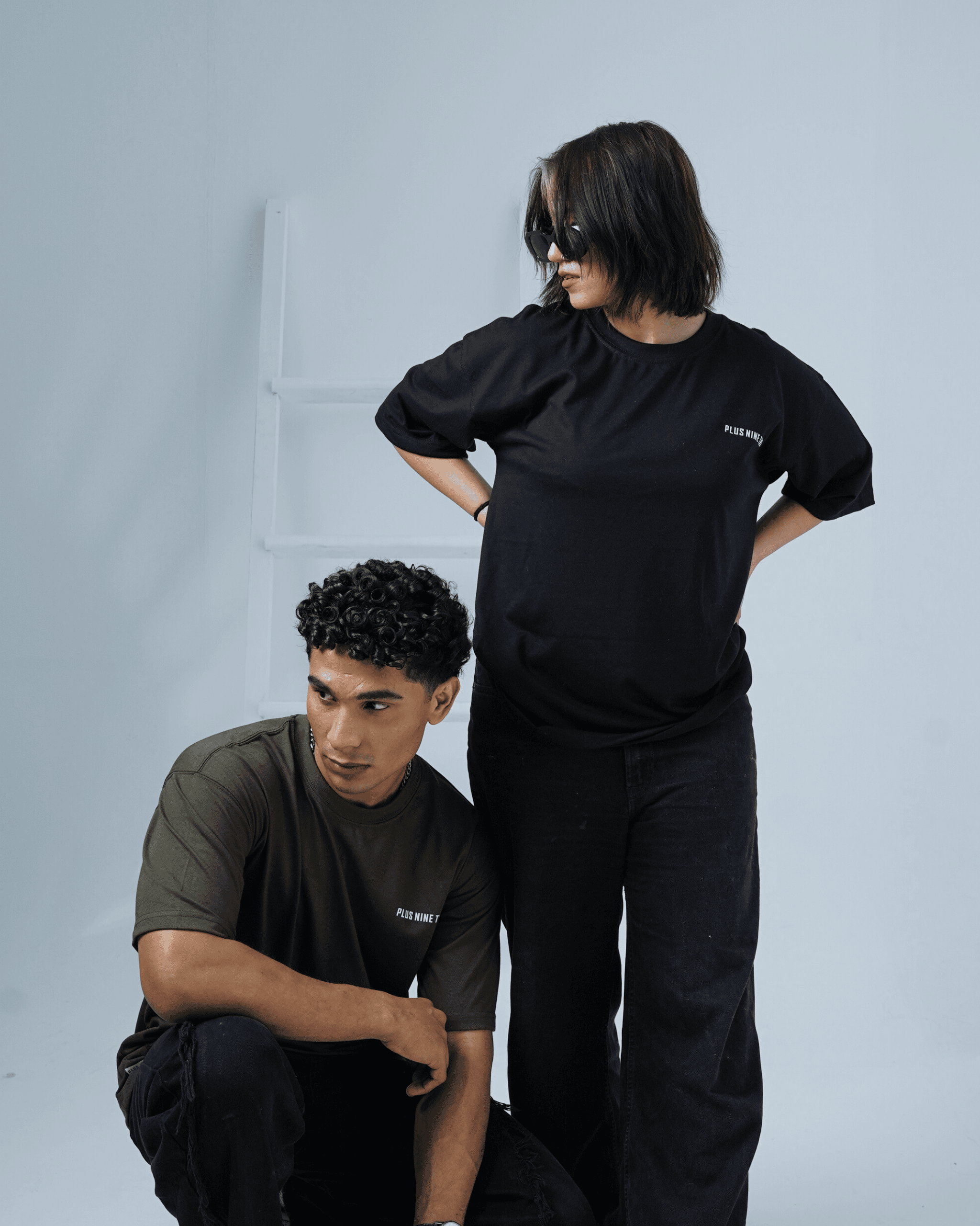Two people a man and woman wearing black and army green oversized t-shirts with a 'PLUS NINE TWO' text against a plain background