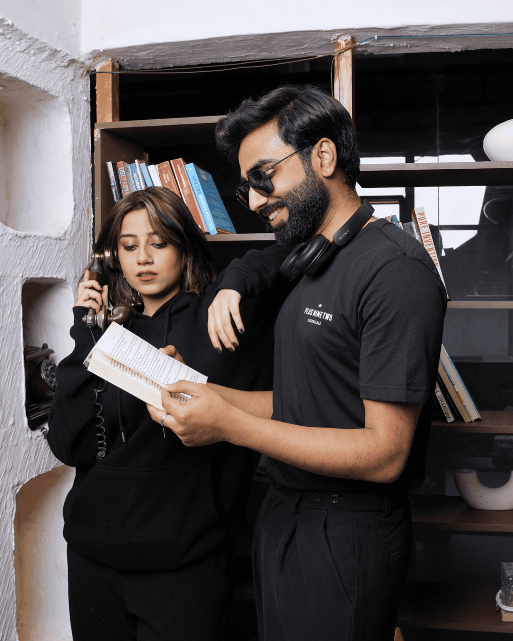 Two people in a room with shelves and books, one holding a book and the other looking at it. The man wearing a black t-shirt and the woman wearing a founders hoodie