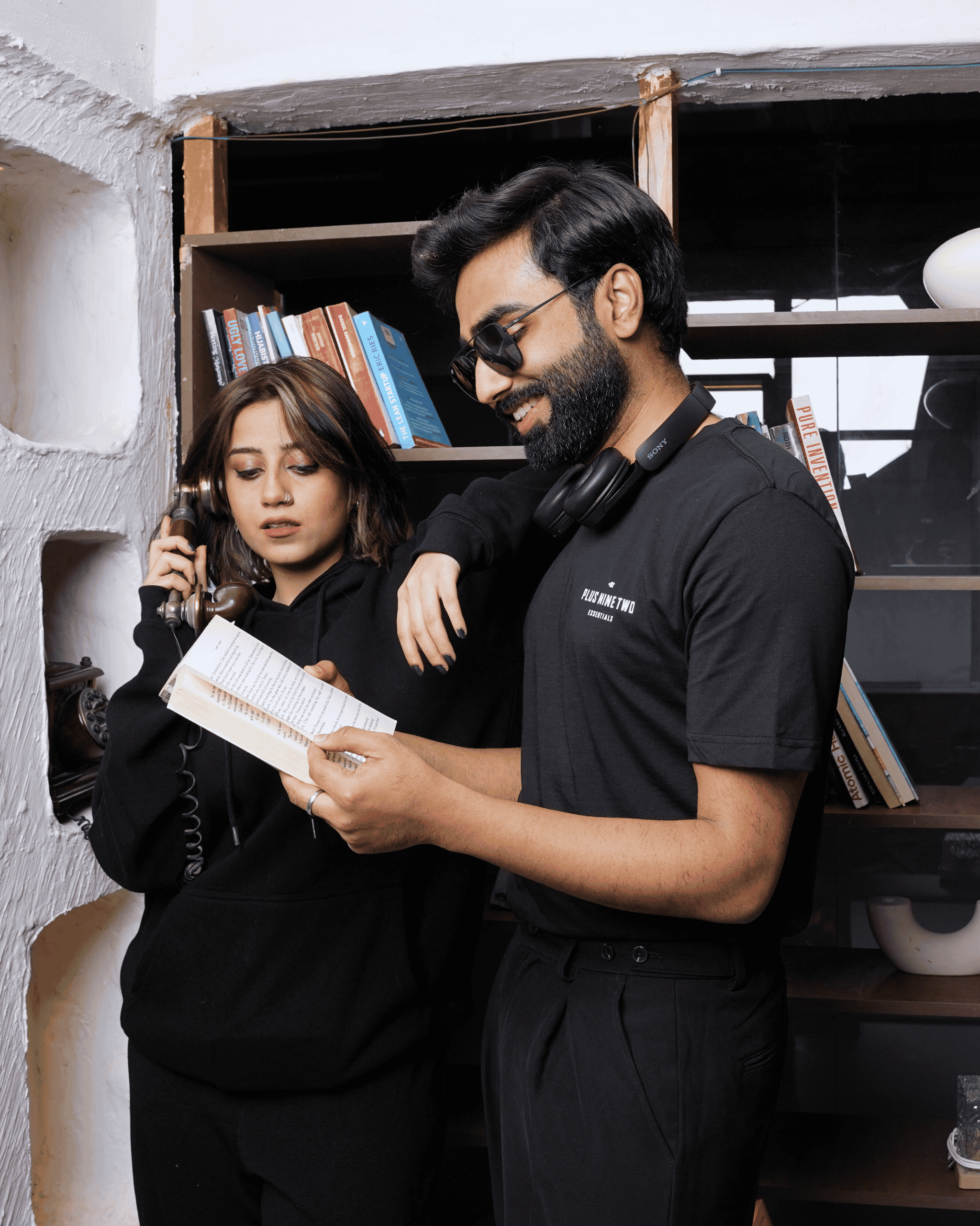Two people in a room with shelves and books, one holding a book and the other looking at it. The man wearing a black t-shirt and the woman wearing a founders hoodie