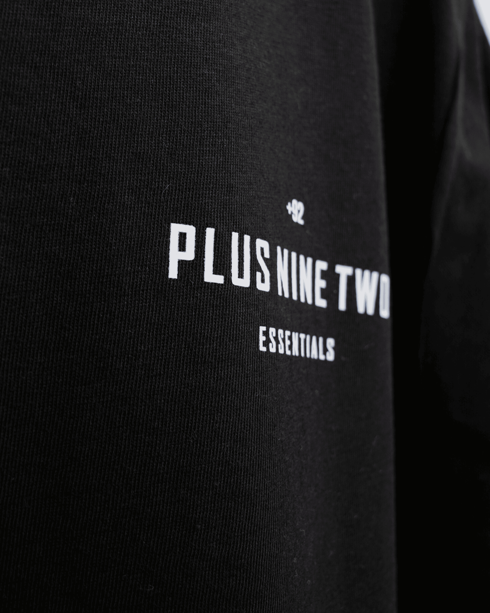 Black t-shirt zoomed into design with '+92' 'PLUS NINE TWO' 'ESSENTIALS' text