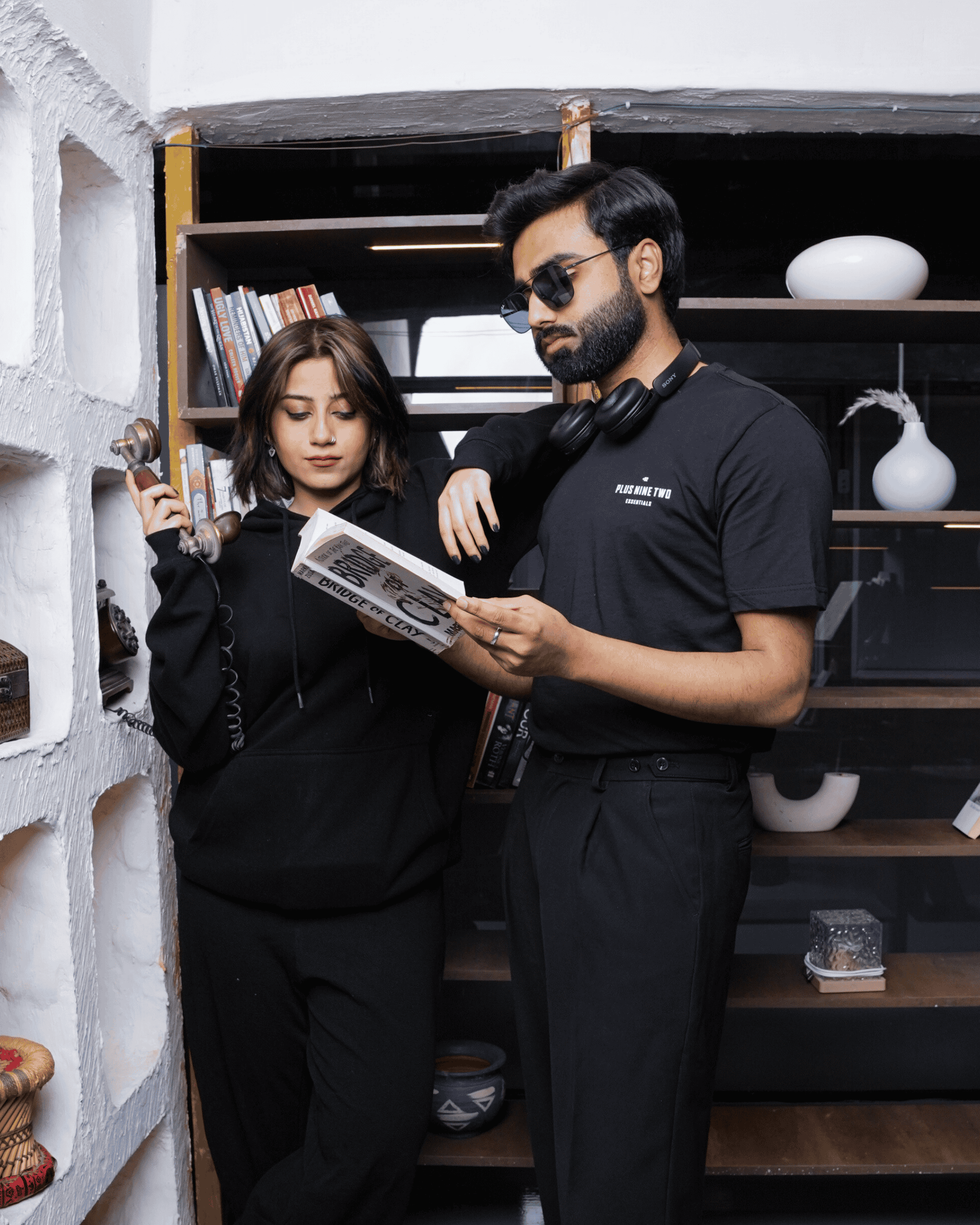 Two people in +92clothing outfits standing in a room with shelves and decor.