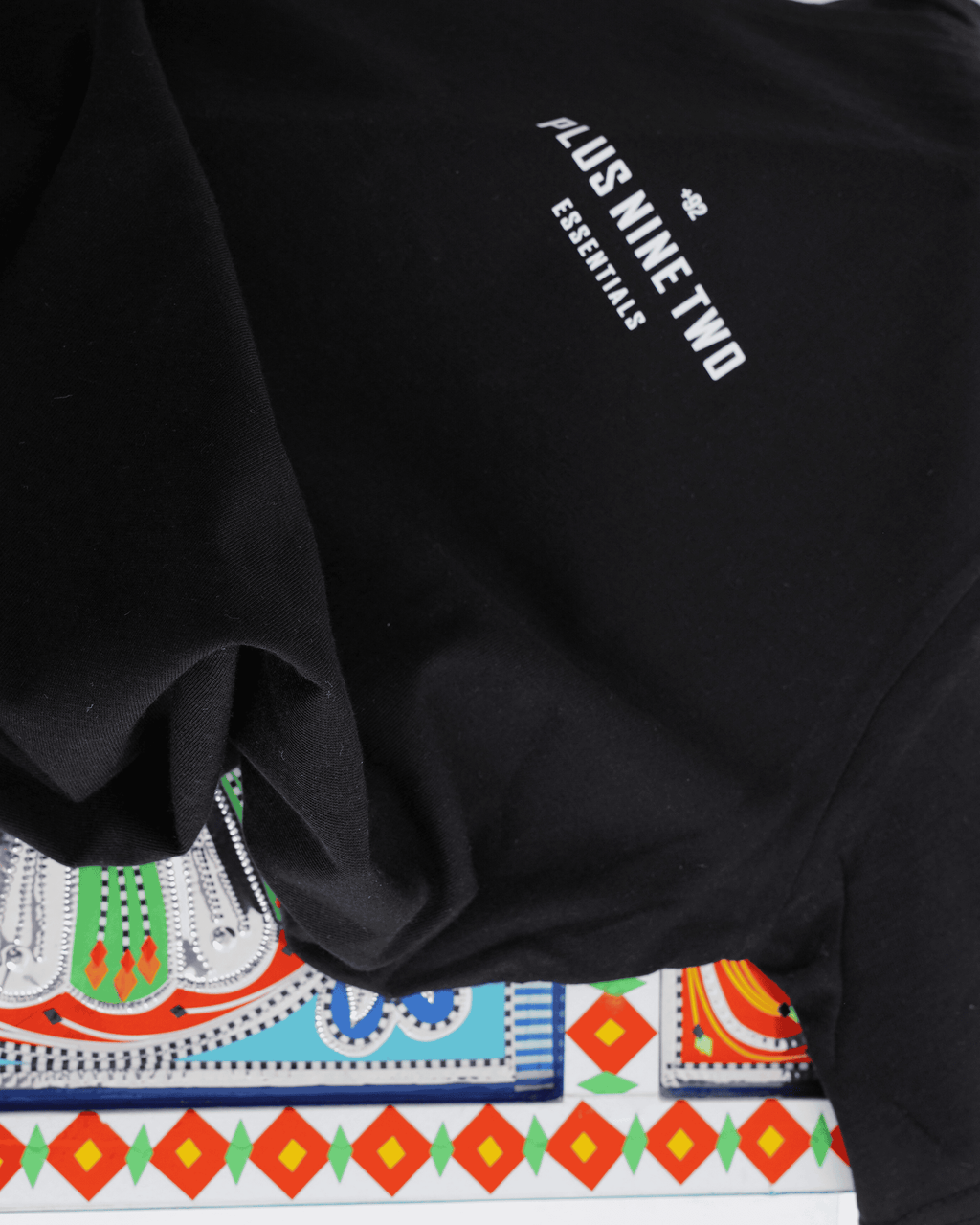 Black t-shirt with text '+92', 'PLUS NINE TWO', 'ESSENTIALS' on a colorful patterned surface