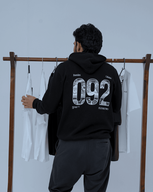 Man wearing a black hoodie with '092' design, standing in front of a rack with clothes.