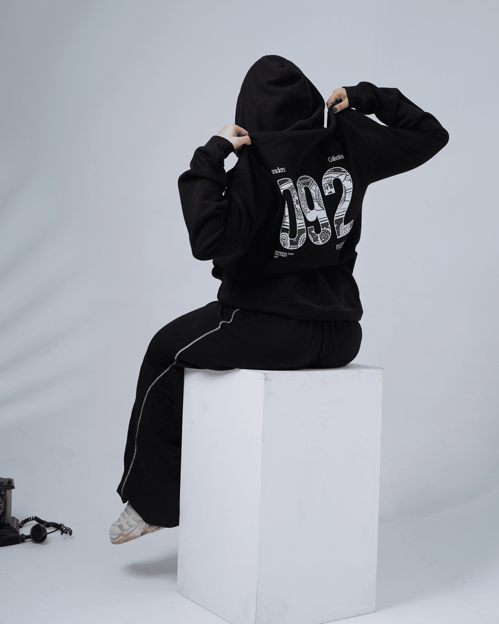 Person wearing a black hoodie with '092' design sitting on a white cube against a gray background