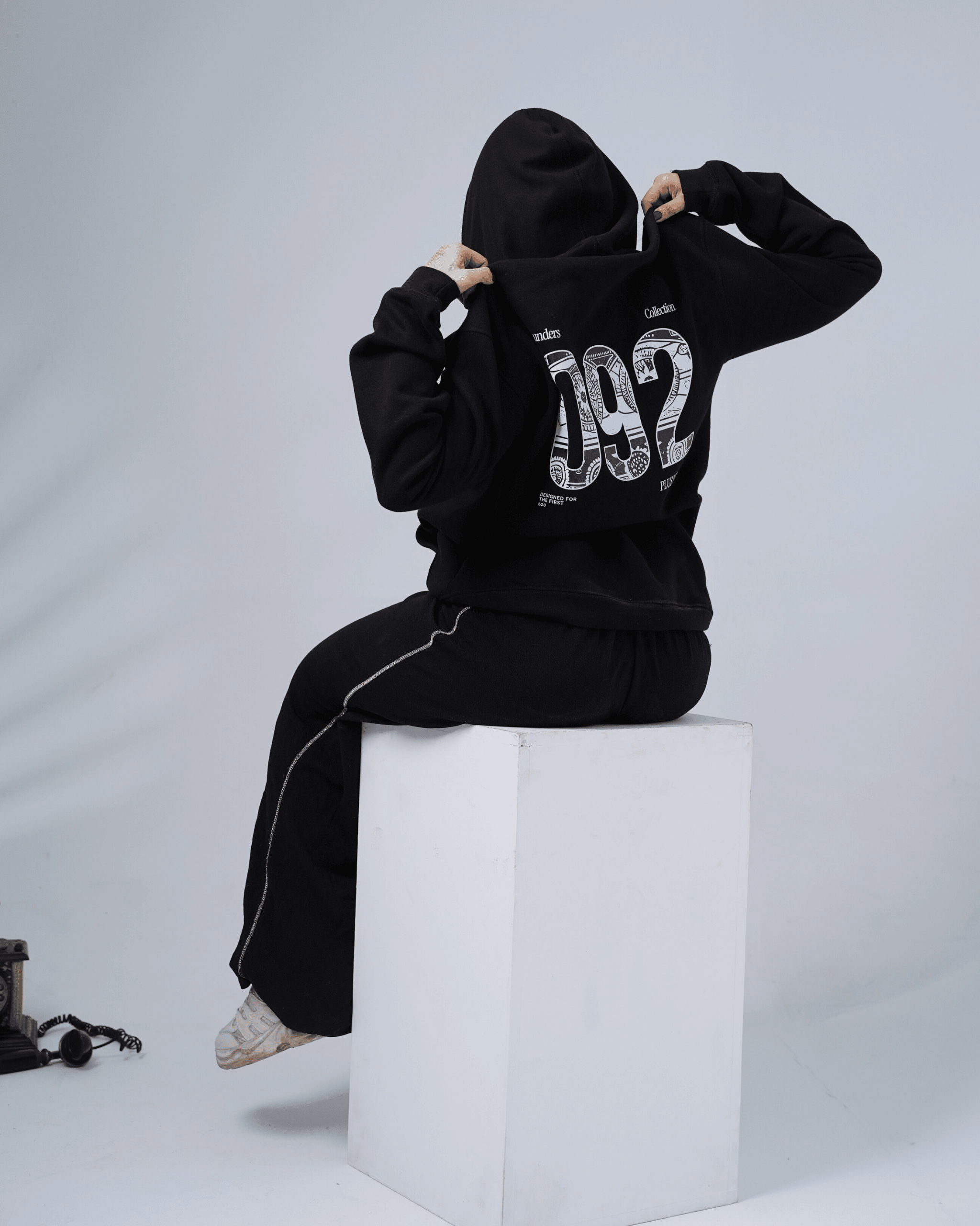 Person wearing a black hoodie with '092' design sitting on a white cube against a gray background