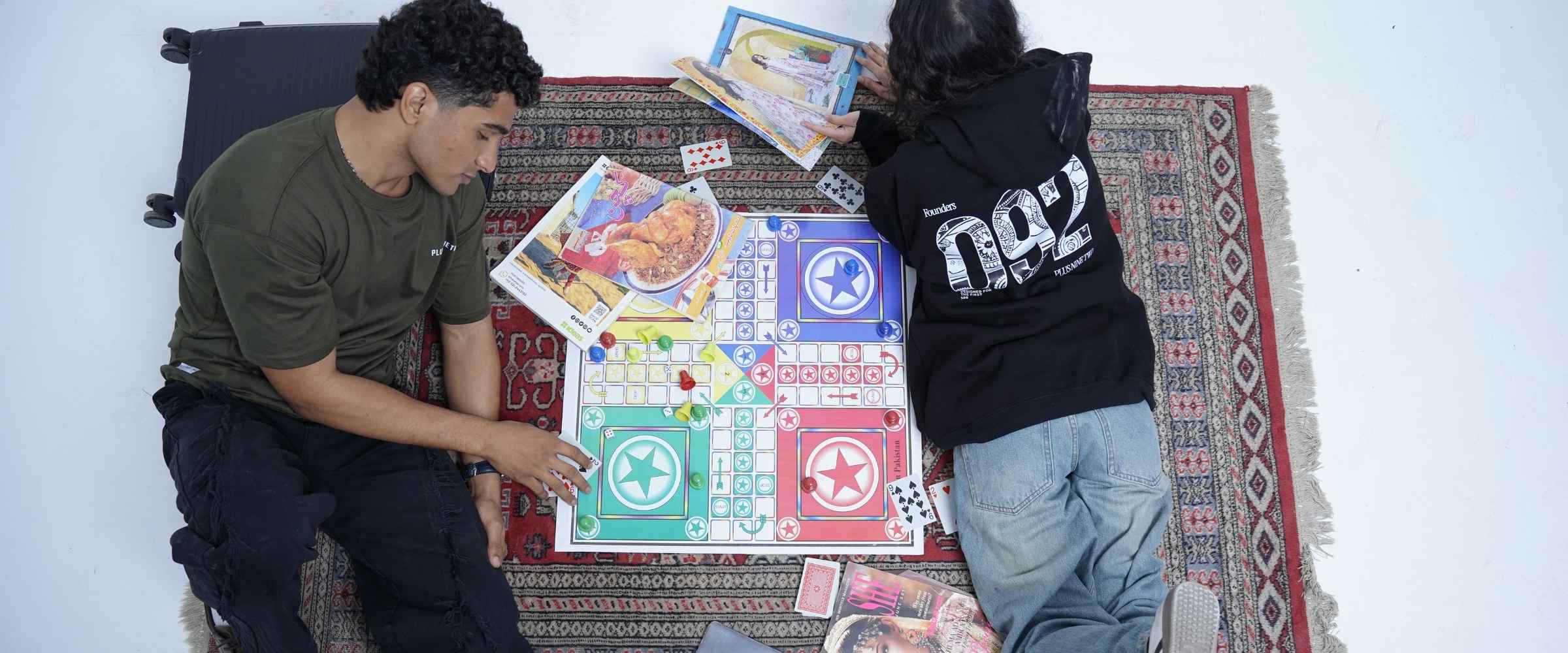 Two people playing a board game on a patterned rug. Man wearing Army Green Essentials Dropshirt and the woman wearing The Founders Hoodie lying on her stomach.