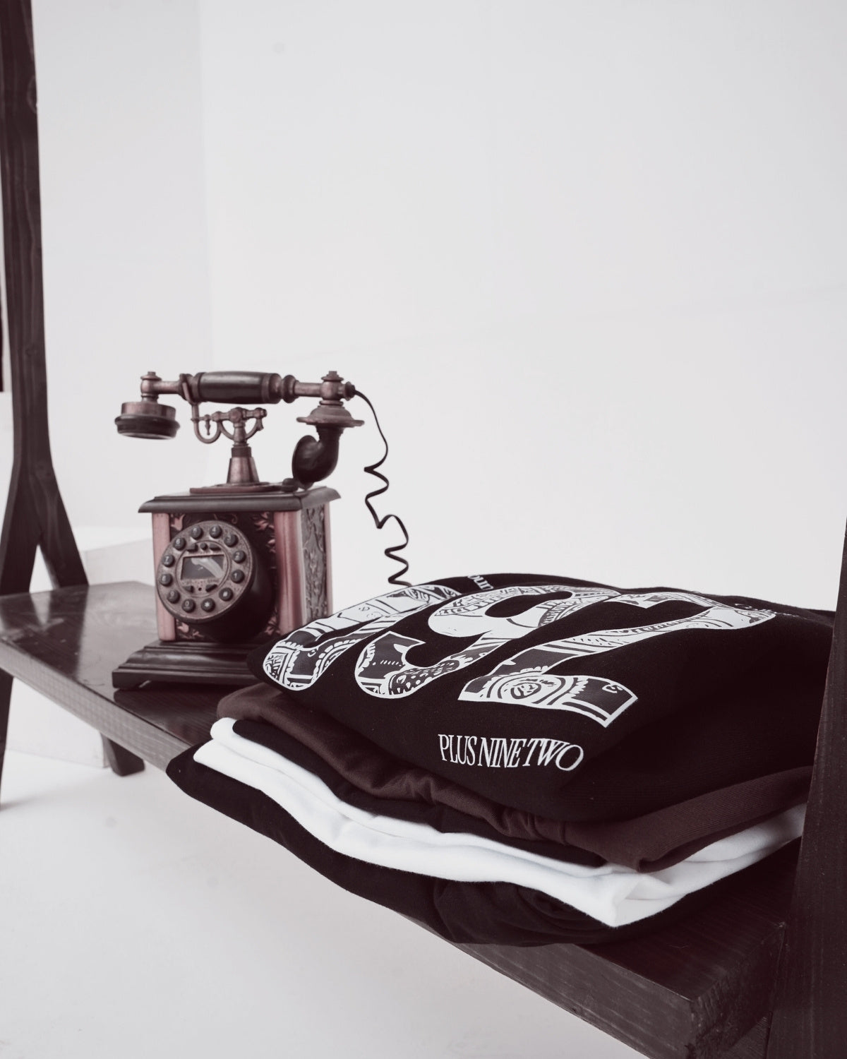 Stack of t-shirts with The founders hoodie folded at the top placed on  wooden shelf next to an old-fashioned telephone.