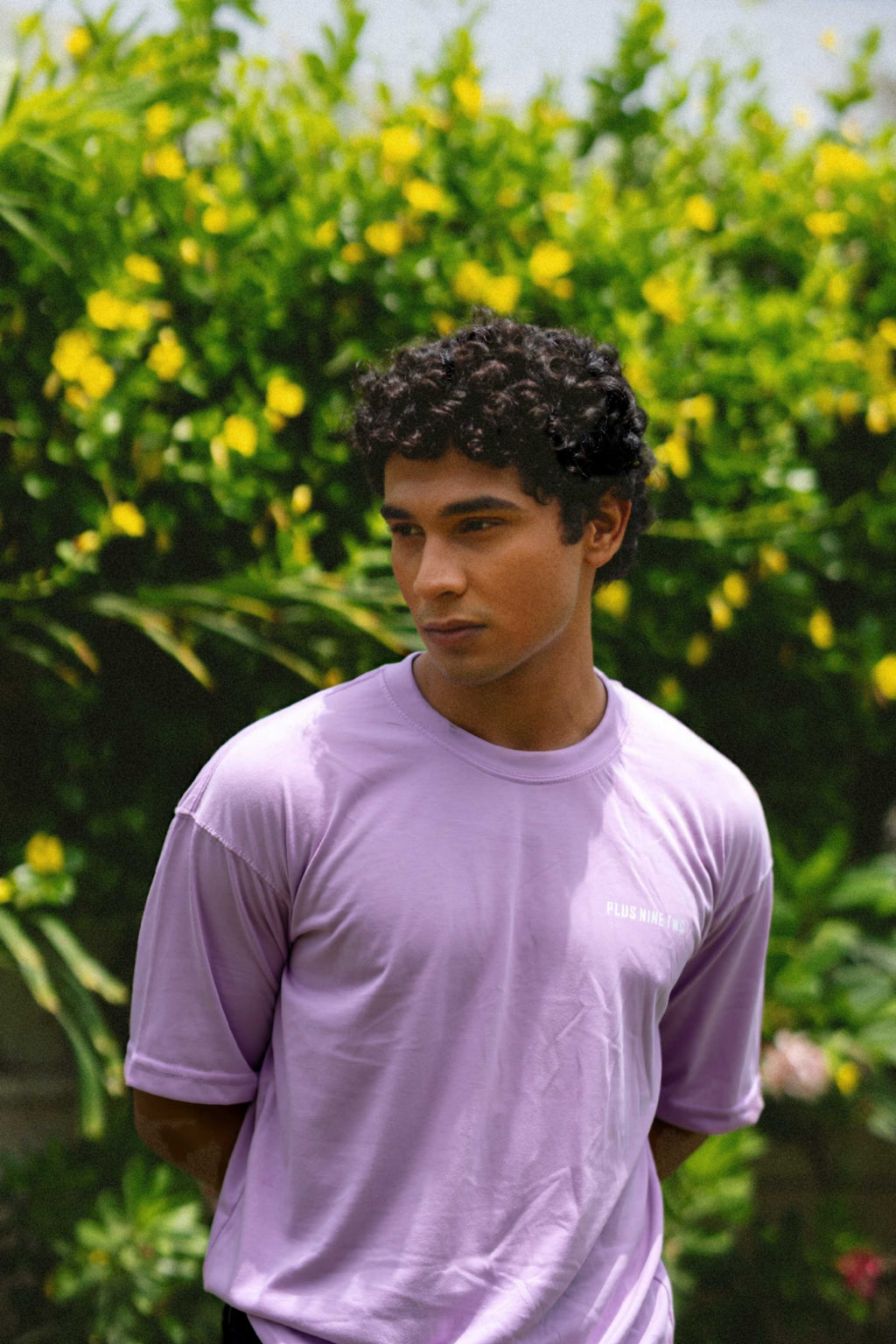 A male model looking down and away while wearing the oversized Lahore Lilac Men's Essentials Drop Shirt in front of a lush garden wall.