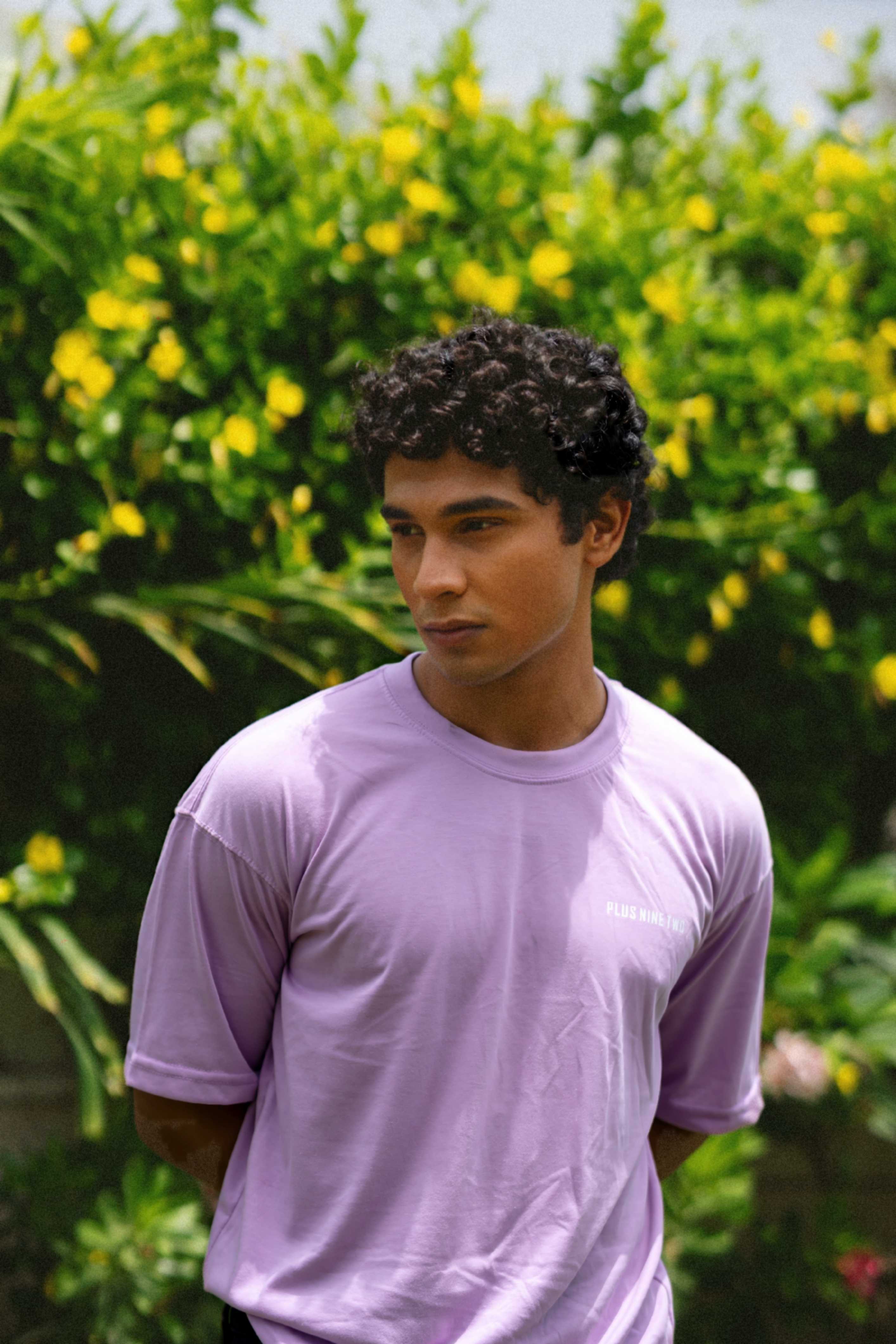 A male model looking down and away while wearing the oversized Lahore Lilac Men's Essentials Drop Shirt in front of a lush garden wall.