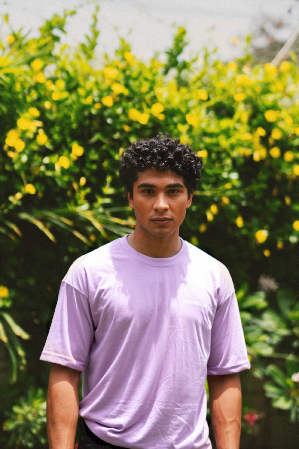 Front view of a male model with curly hair wearing the Lahore Lilac Men's Essentials Drop Shirt against a vibrant green and yellow foliage background.