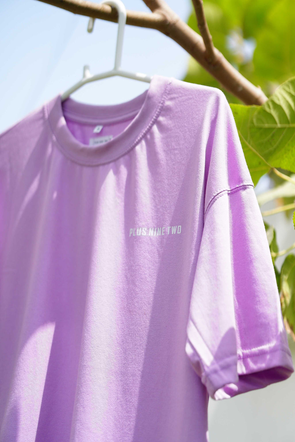 Angled close-up of the Lahore Lilac Men's Essentials Drop Shirt on a hanger, focusing on the crew neck, dropped shoulder seam, and white text logo.