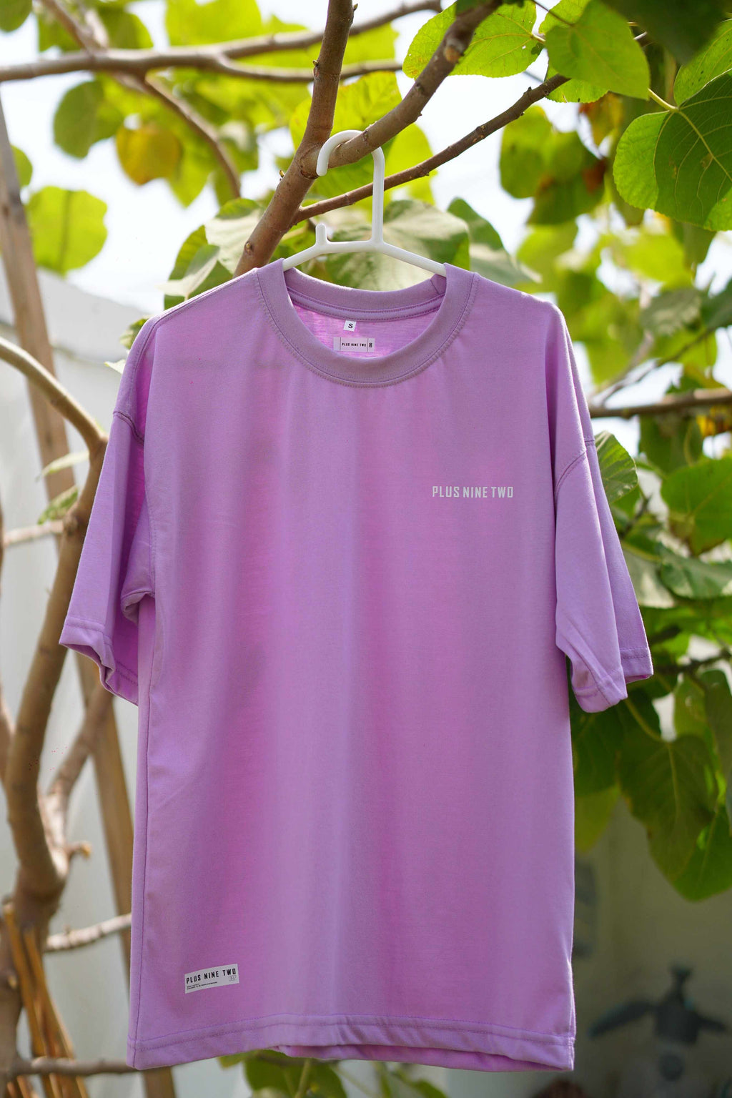 Lahore Lilac Men's Essentials Drop Shirt hanging on a white hanger outdoors, showcasing its oversized fit, crew neckline, and subtle chest branding.