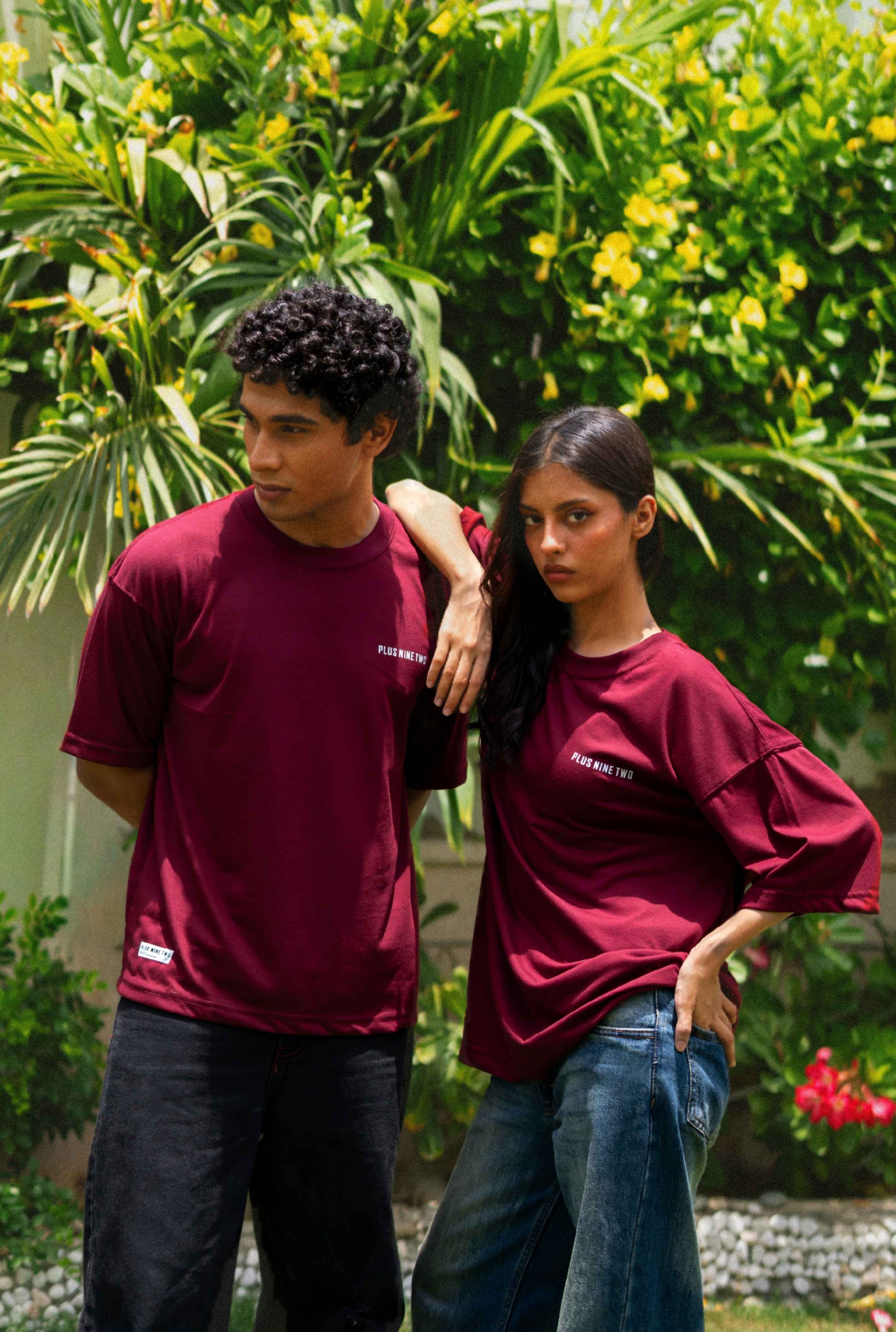 Two models wearing matching Ajrak Maroon Men's Essentials Drop Shirt with white text on the chest, standing outdoors in front of lush green foliage.
