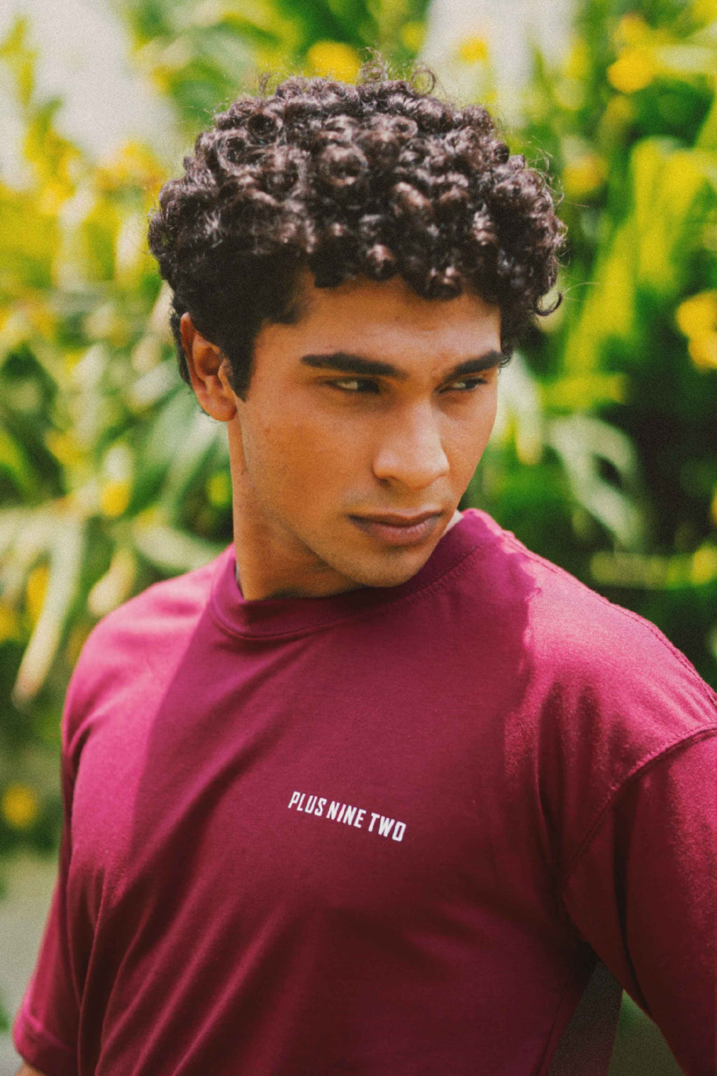 A man with curly dark hair wearing Ajrak Maroon Men's Essentials Drop Shirt with 'PLUS NINE TWO' printed in white on the chest, set against a blurred green and yellow outdoor background.