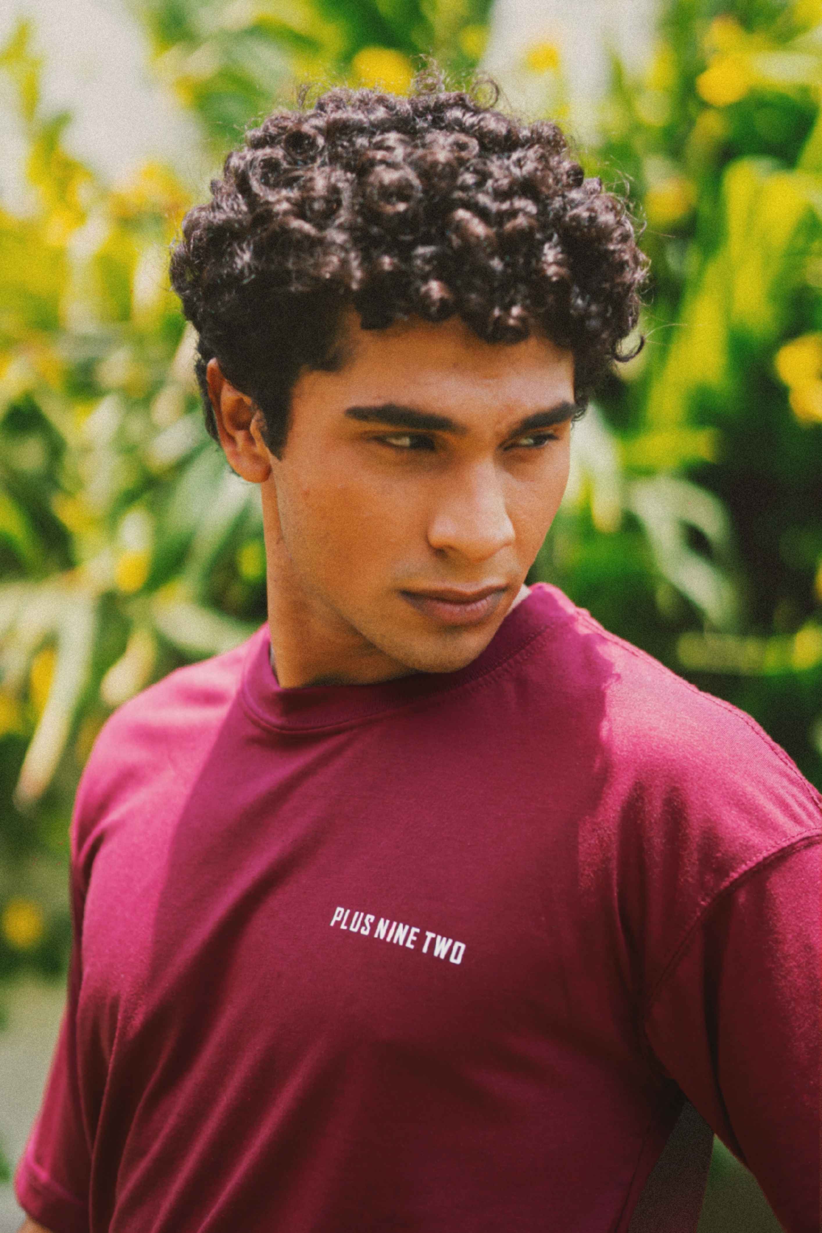 A man with curly dark hair wearing Ajrak Maroon Men's Essentials Drop Shirt with 'PLUS NINE TWO' printed in white on the chest, set against a blurred green and yellow outdoor background.