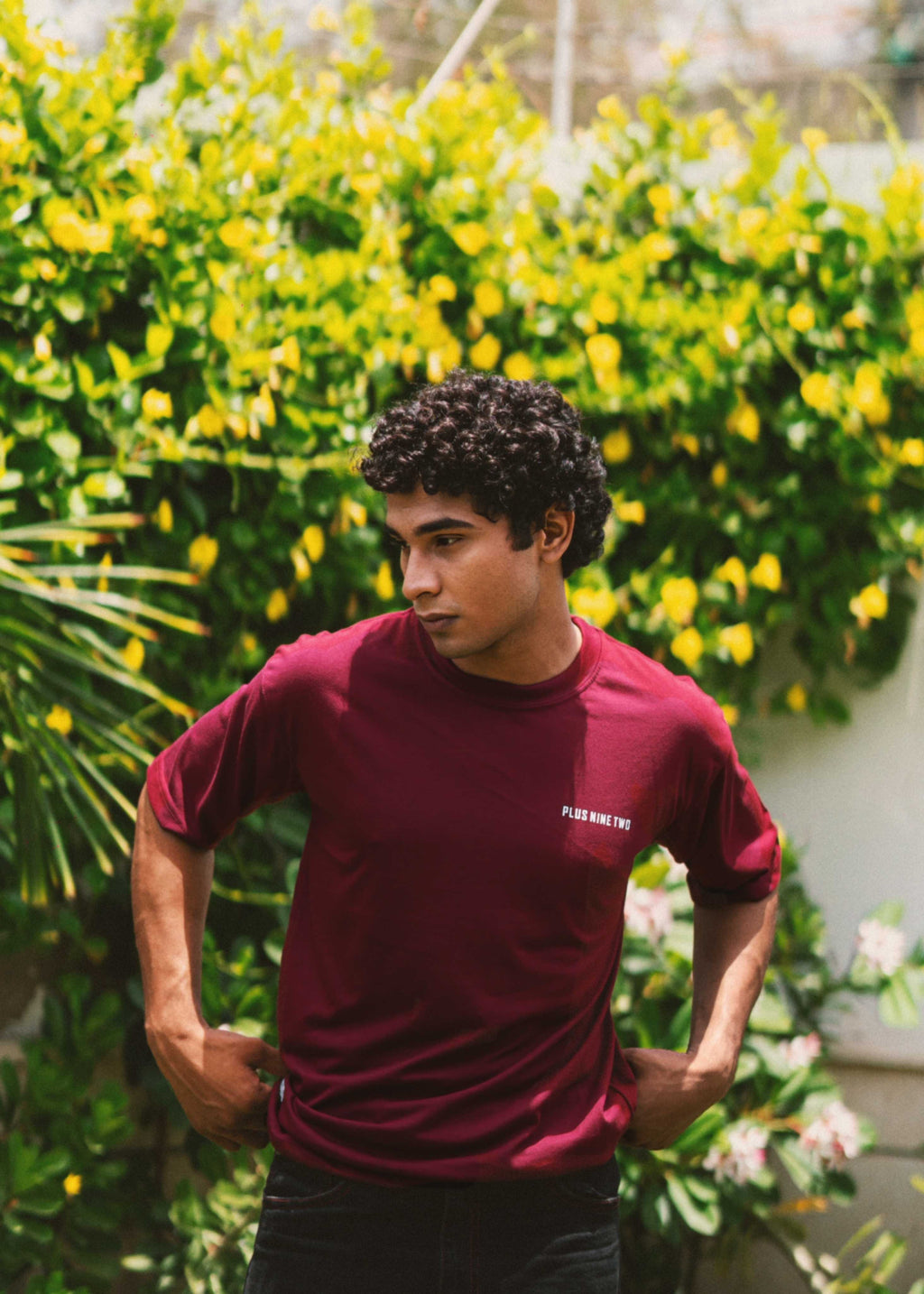 Male model looking down to the side while wearing the streetwear-style Ajrak Maroon Men's Essentials Drop Shirt in a garden setting.