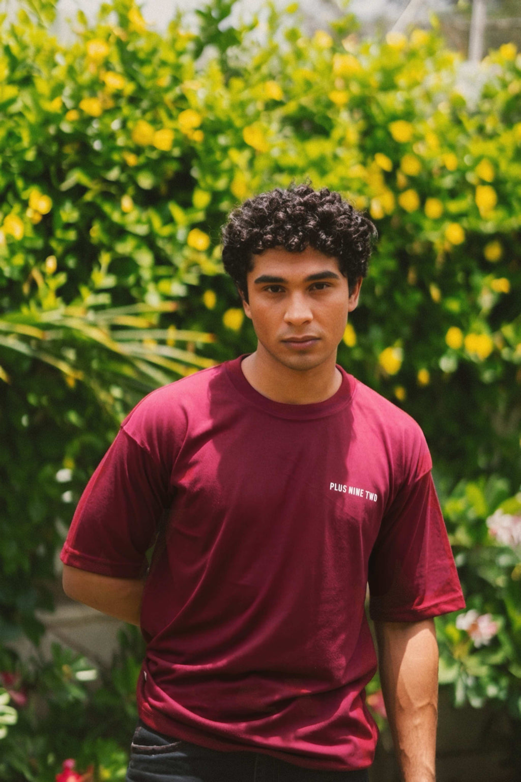 Front view of a male model with curly hair modeling the relaxed-fit Ajrak Maroon Men's Essentials Drop Shirt against a backdrop of yellow flowers.