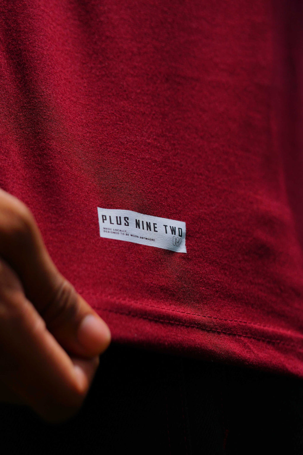 Extreme close-up of a hand holding the bottom hem of the maroon cotton t-shirt, highlighting the "PLUS NINE TWO" woven branding tag.