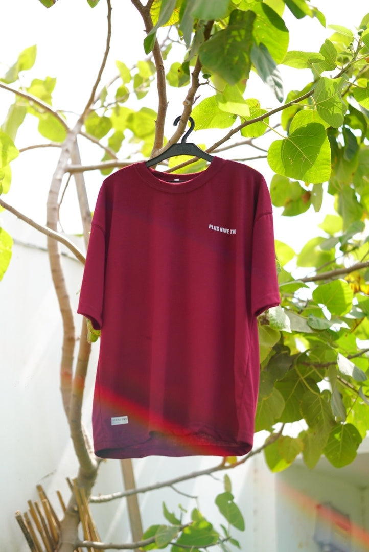 Full front view of the oversized Ajrak Maroon Men's Essentials Drop Shirt hanging on a black hanger outdoors against green foliage.