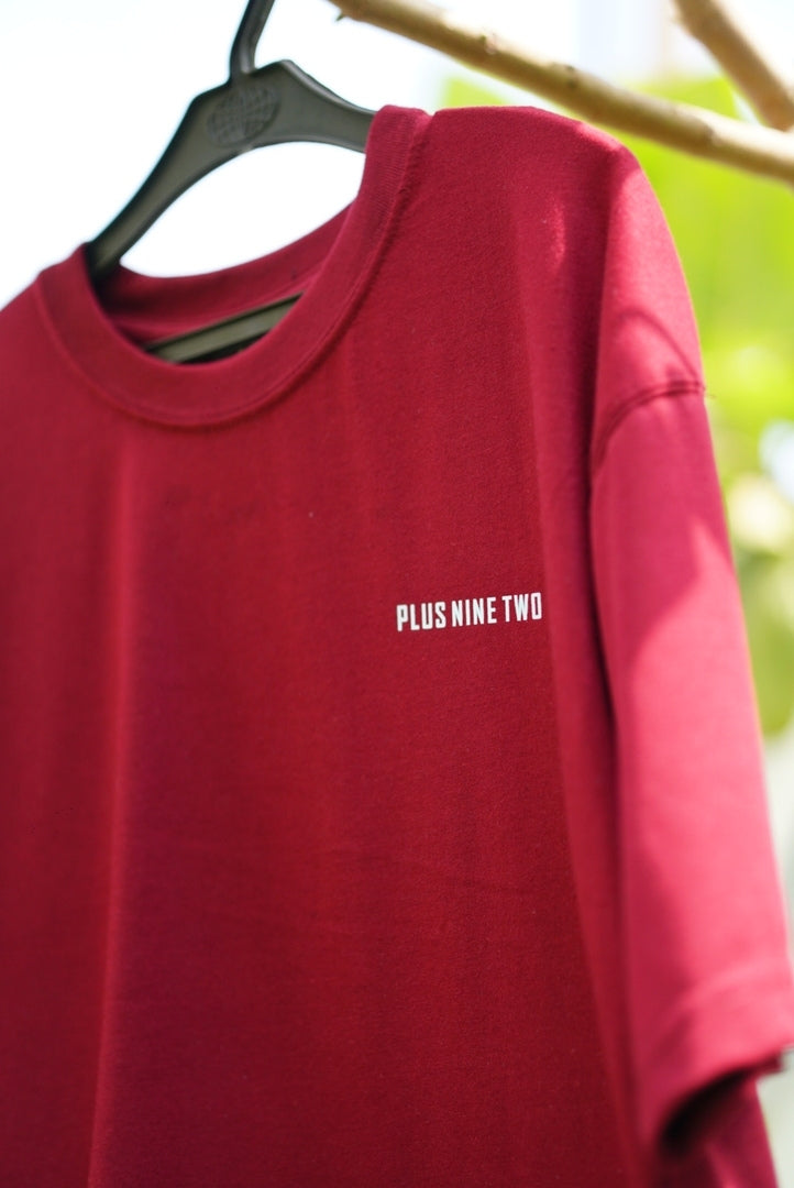 Ajrak Maroon Women's Essentials Drop Shirt hanging on a black hanger, featuring the text 'PLUS NINE TWO' printed in white on the left chest. The background is a soft-focus outdoor scene with green foliage and a wooden branch.