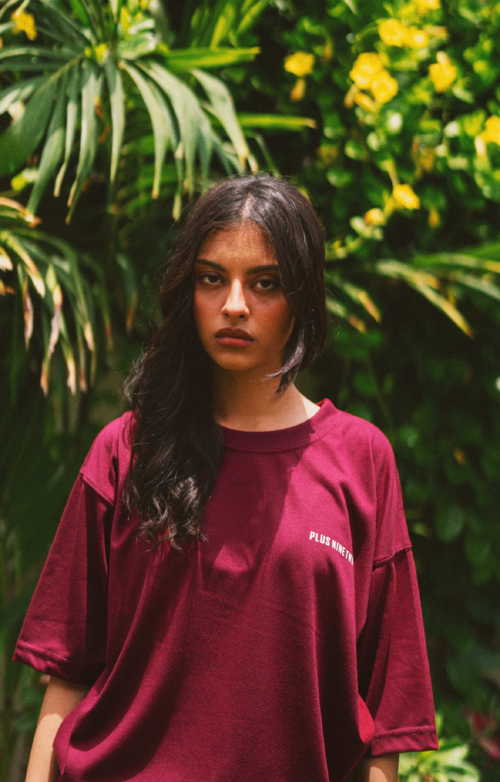 A woman with long dark hair wearing Ajrak Maroon Women's Essentials Drop Shirt with white text on the chest, standing in front of a lush green background with yellow flowers.
