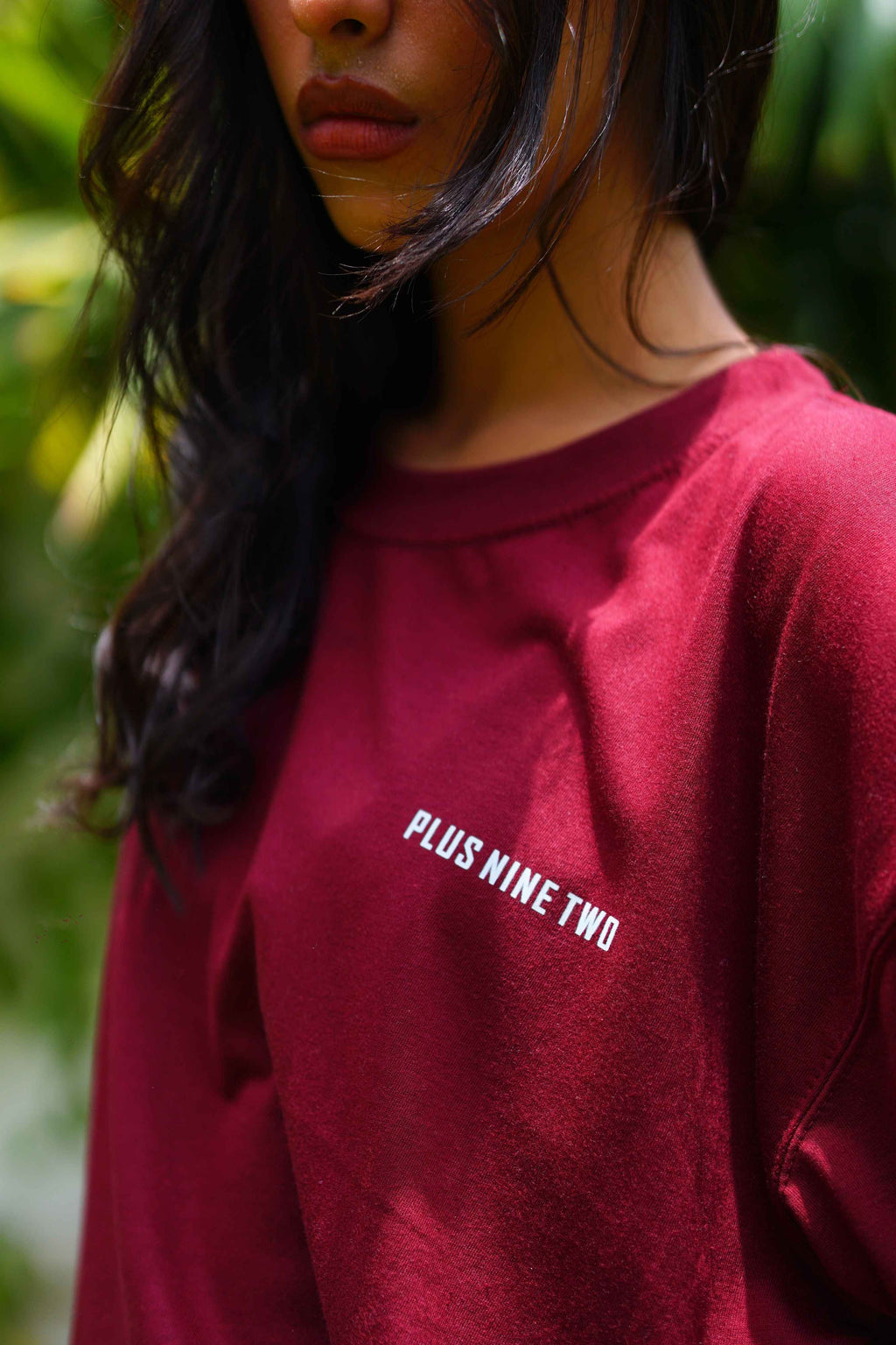 A close-up of a person wearing Ajrak Maroon Women's Essentials Drop Shirt with 'PLUS NINE TWO' printed in white text on the chest, set against a blurred green background.