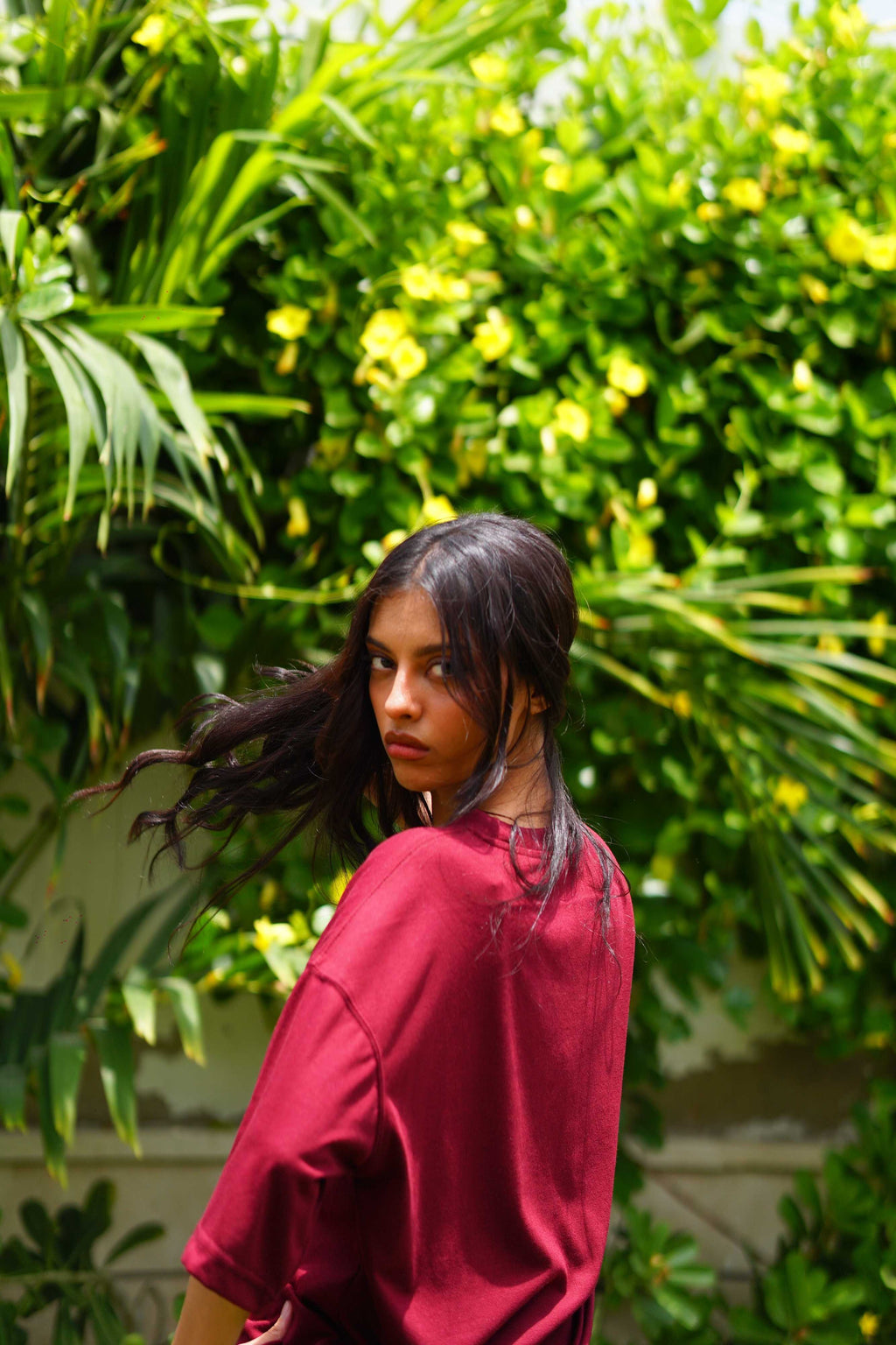 A woman with long dark hair wearing Ajrak Maroon Women's Essentials Drop Shirt, looking back over her shoulder against a lush green tropical foliage background with yellow flowers.