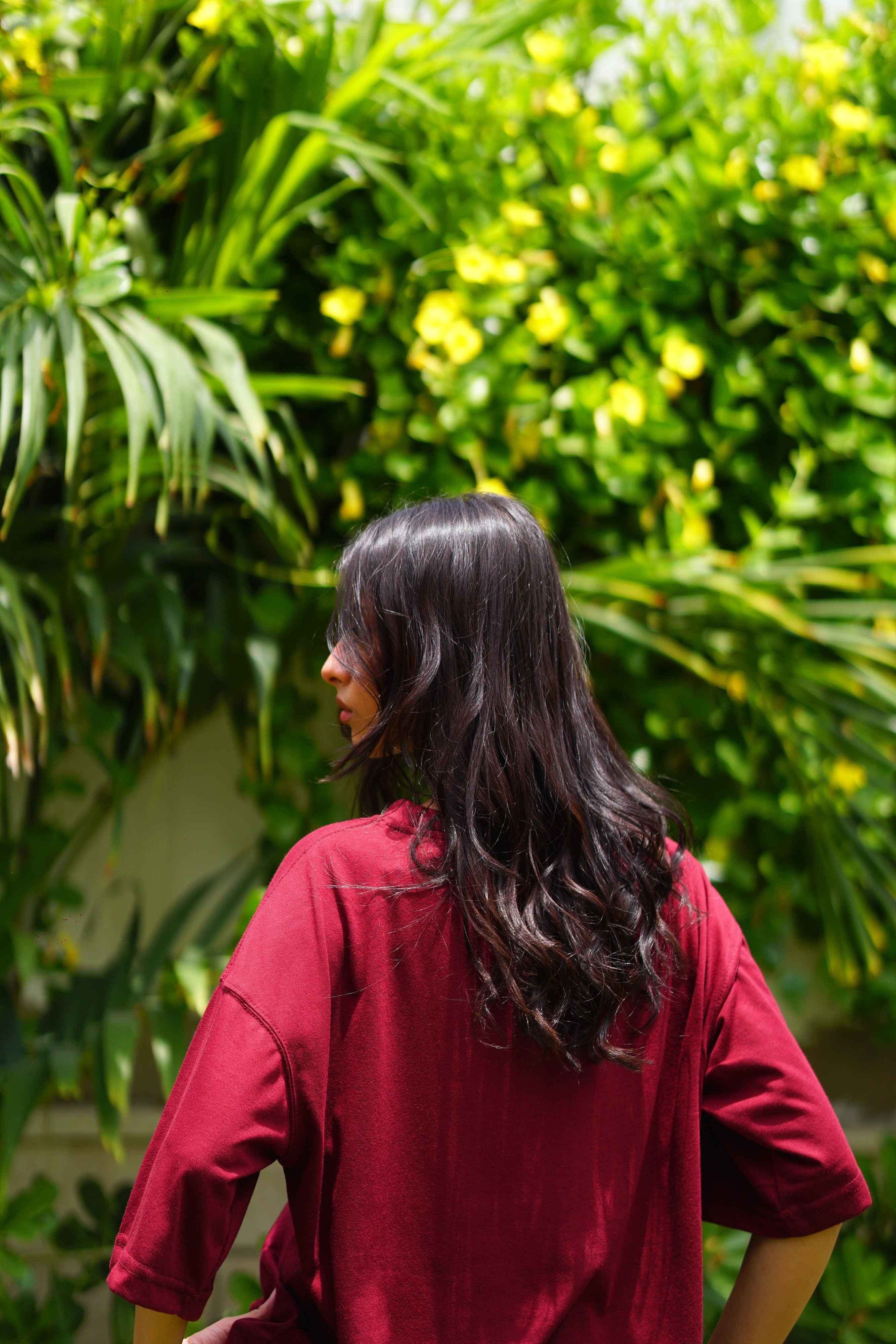 A woman with long dark hair, seen from behind, wearing an Ajrak Maroon Women's Essentials Drop Shirt, standing in front of a lush green garden wall with yellow flowers.