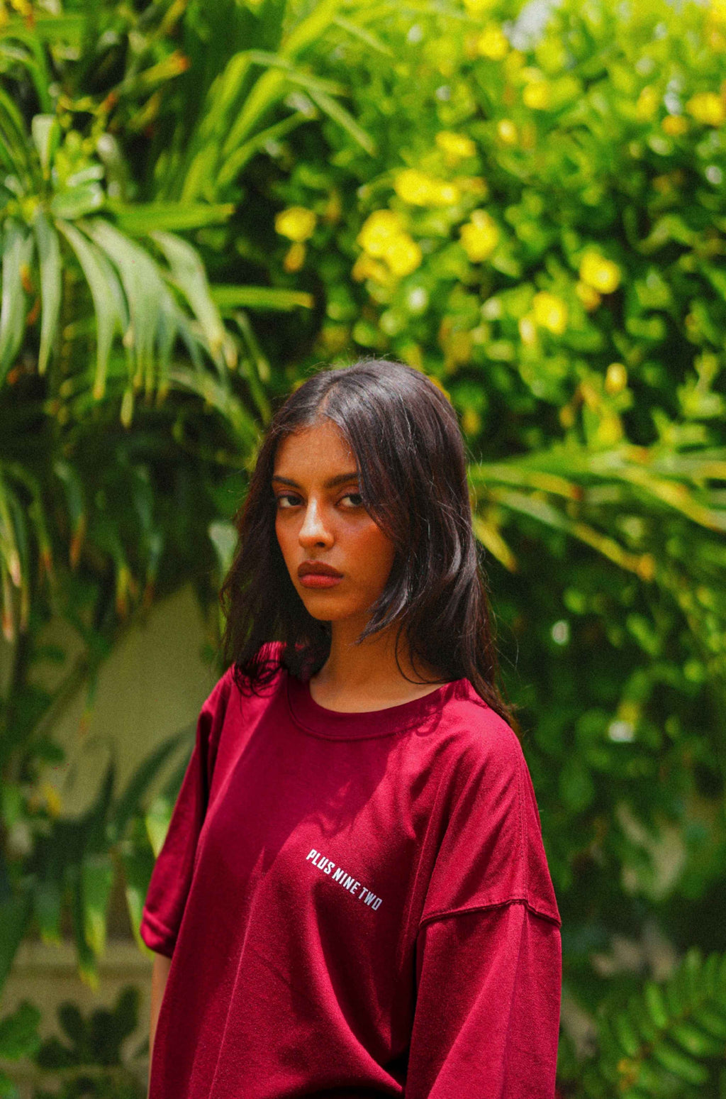 A woman with dark hair wearing Ajrak Maroon Women's Essentials Drop Shirt with white text on the chest, standing in front of a lush green foliage background with yellow flowers.