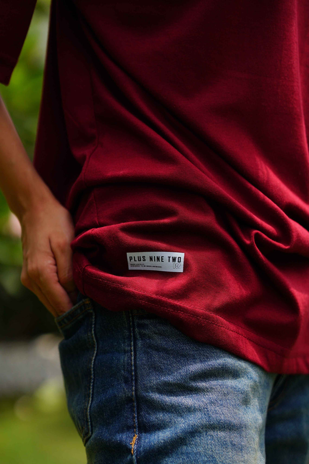 A close-up of a person wearing Ajrak Maroon Women's Essentials Drop Shirt and blue jeans, showing a white brand label on the bottom hem of the shirt that reads 'PLUS NINE TWO'.