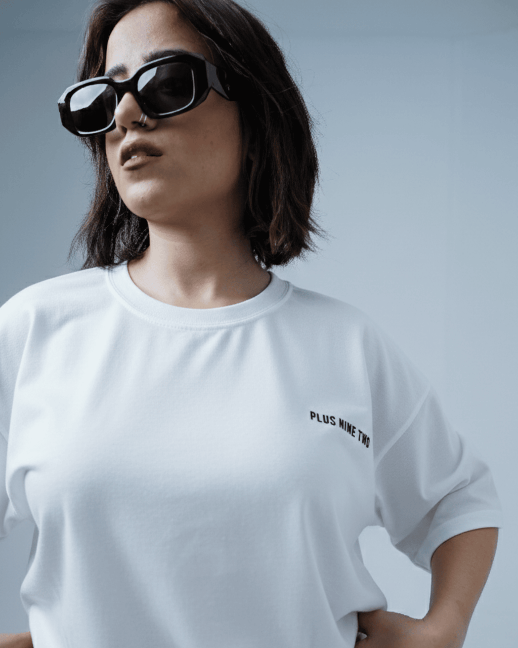 Woman wearing a white oversized t-shirt with text 'PLUS NINE TWO', standing against a plain background