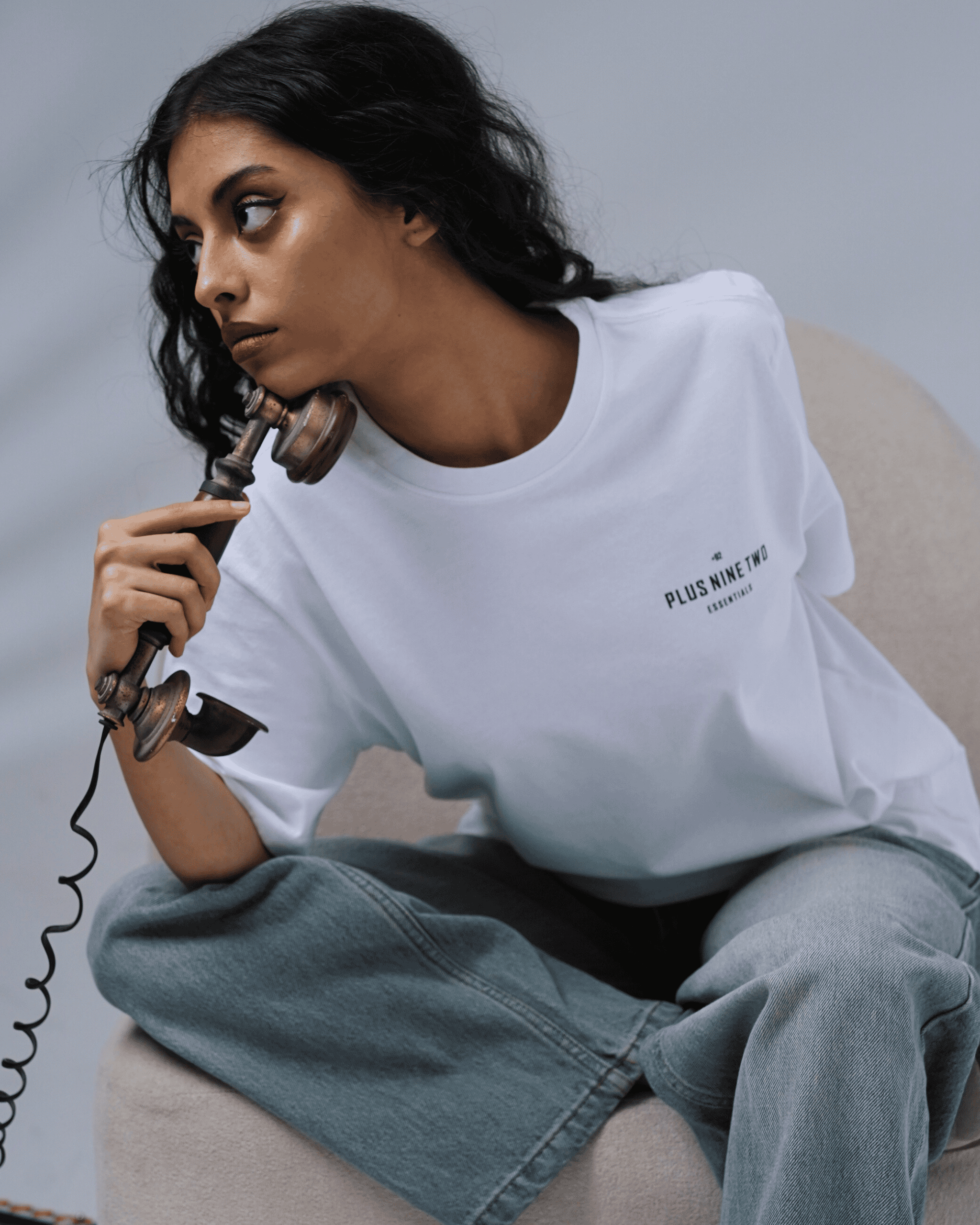 A girl wearing a white t-shirt with text +92/PLUS NINE TWO/ ESSENTIALS, sitting on a light-colored sofa with an antique phone in hand.