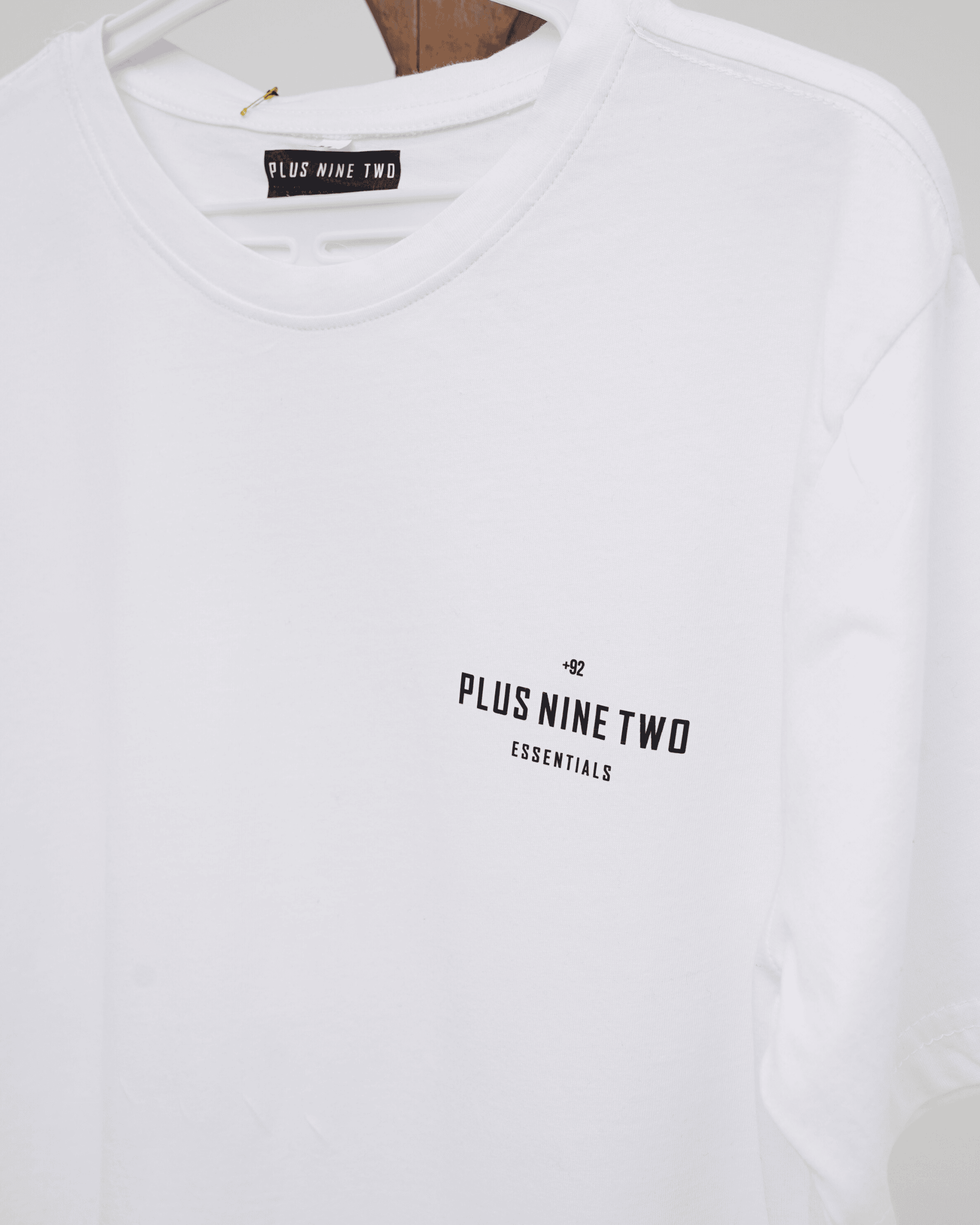 White t-shirt with '+92', 'PLUS NINE TWO' , 'ESSENTIALS' design on a hanger.