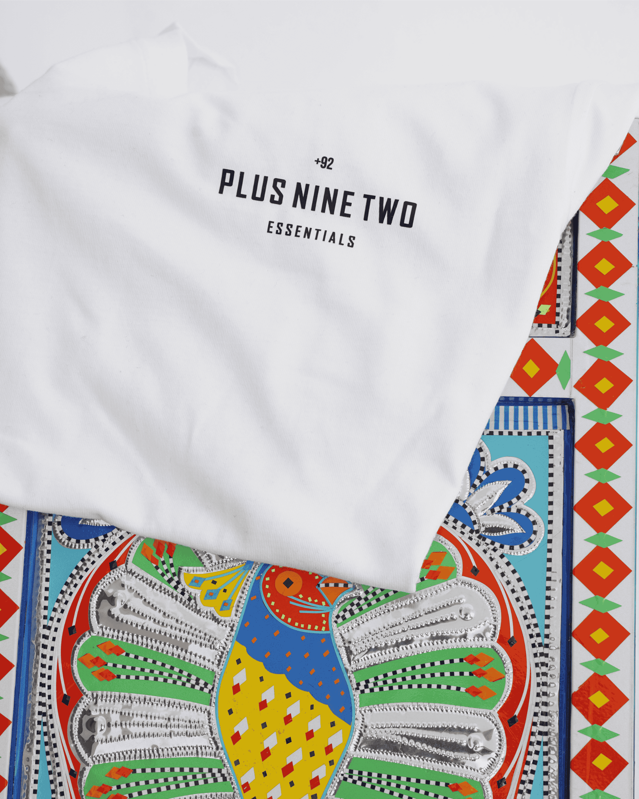 White t-shirt with '+92' 'Plus Nine Two' 'Essentials' text and colorful geometric design on a white background.